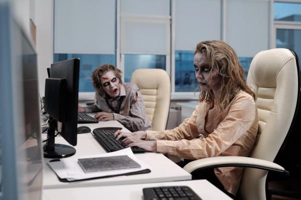 Young zombie businesswoman working at her desk, with a deceased male colleague in the background.